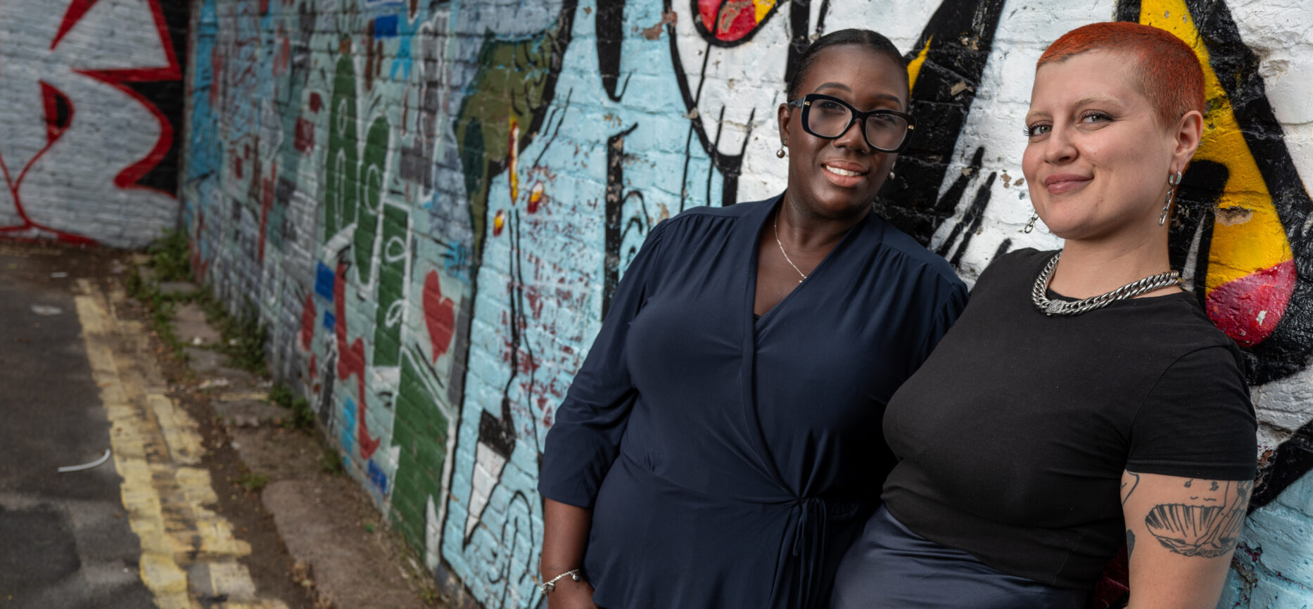 Two women leaning against a wall painted with graffiti art smiling