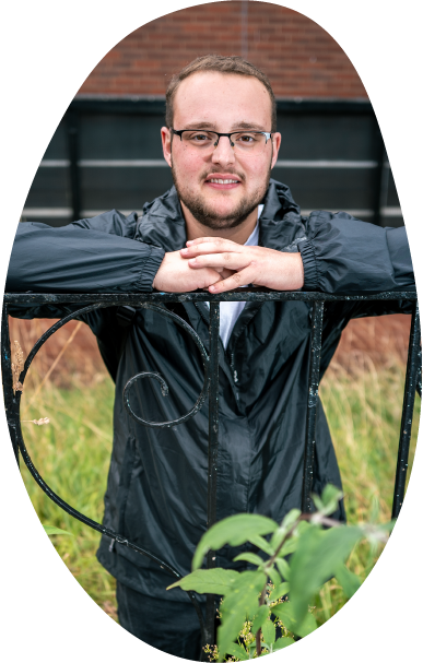 A young man smiling and looking hopeful, wearing glasses and an anorak, leaning on a gate