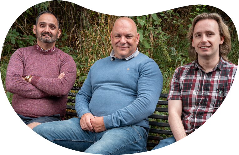 A photo of three men smiling together, sat of a bench in the park