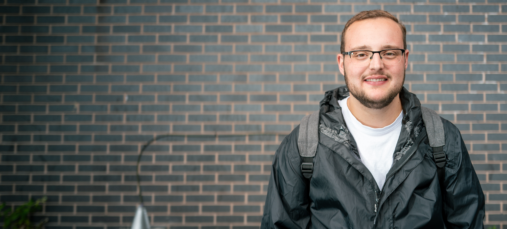 A photo of a young man smiling, wearing a raincoat and standing in front of a grey brick wall outside